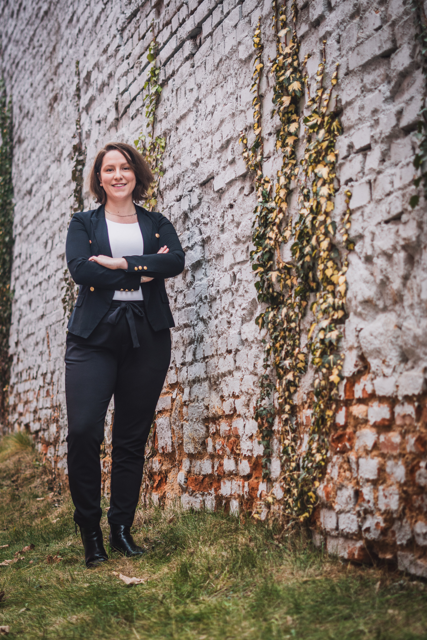 Venkovní business portrét elegantní ženy v černém saku, stojící před starou cihlovou zdí porostlou břečťanem.