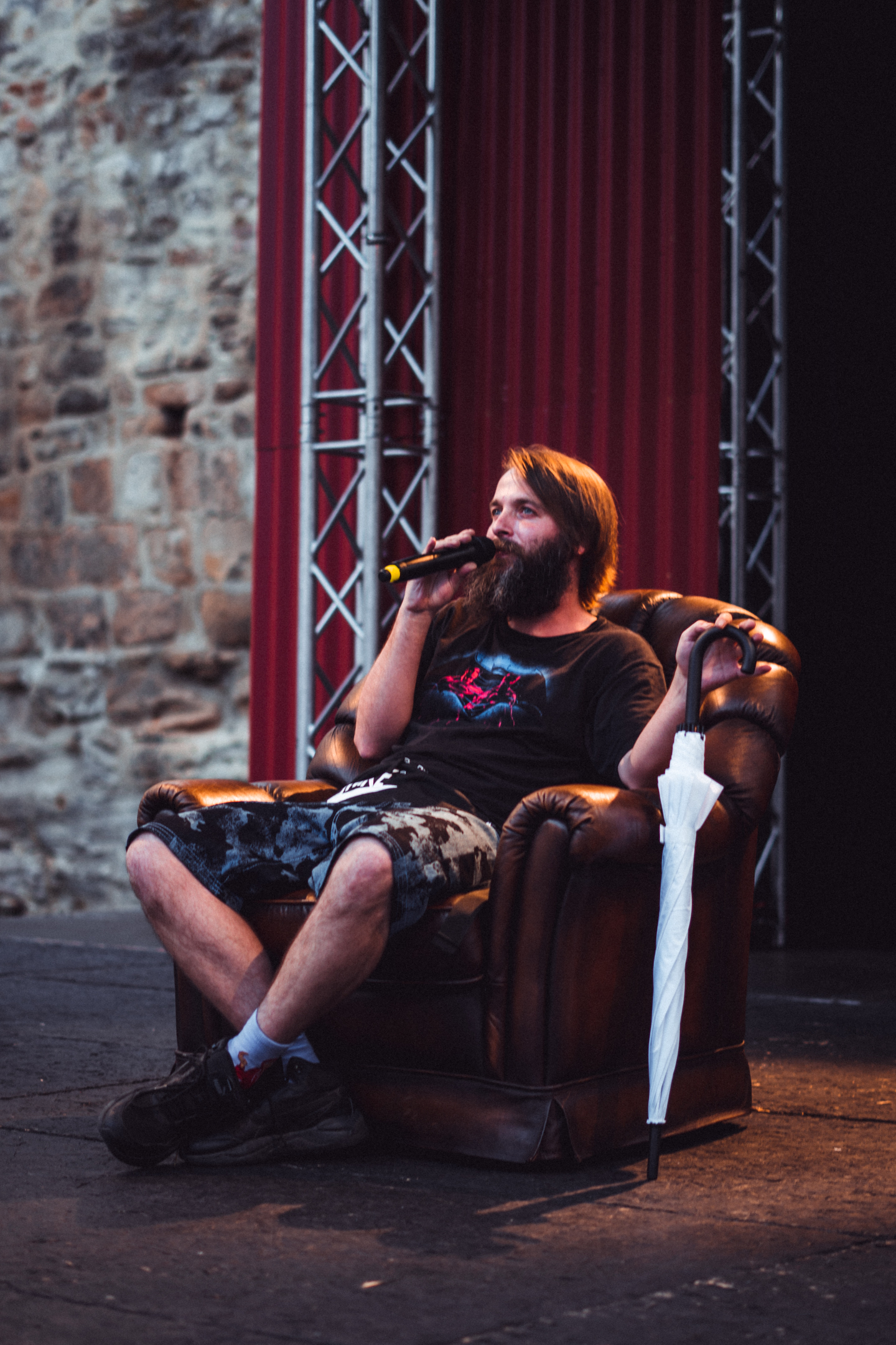 Performer s plnovousem sedí v křesle na pódiu a mluví do mikrofonu během venkovního slam poetry vystoupení.