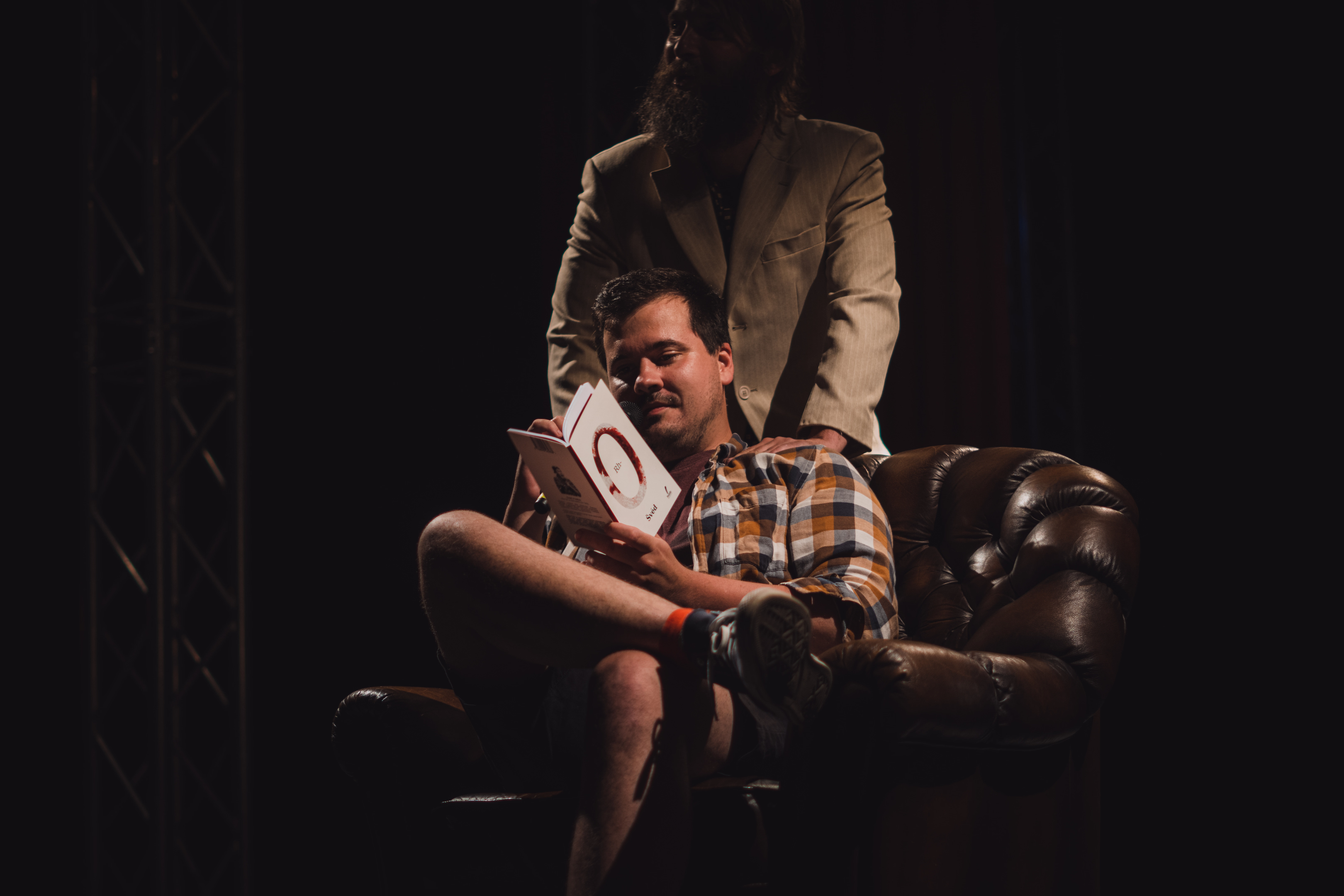 Účinkující sedí v křesle na tmavém jevišti a čte z knihy během divadelního nebo slam poetry vystoupení.