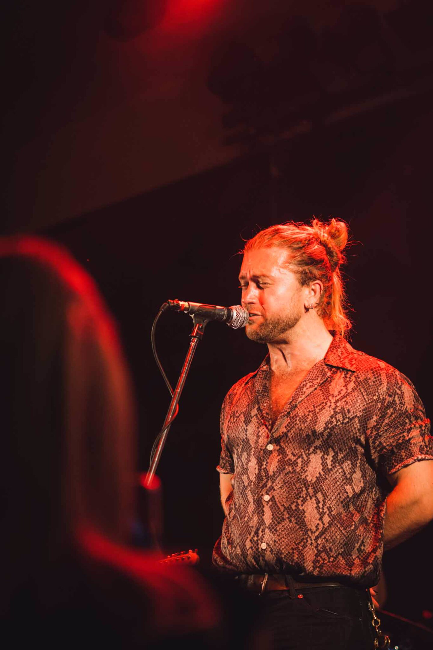 Zpěvák s dlouhými vlasy v culíku zpívá do mikrofonu na pódiu zalitém červeným světlem během koncertu.