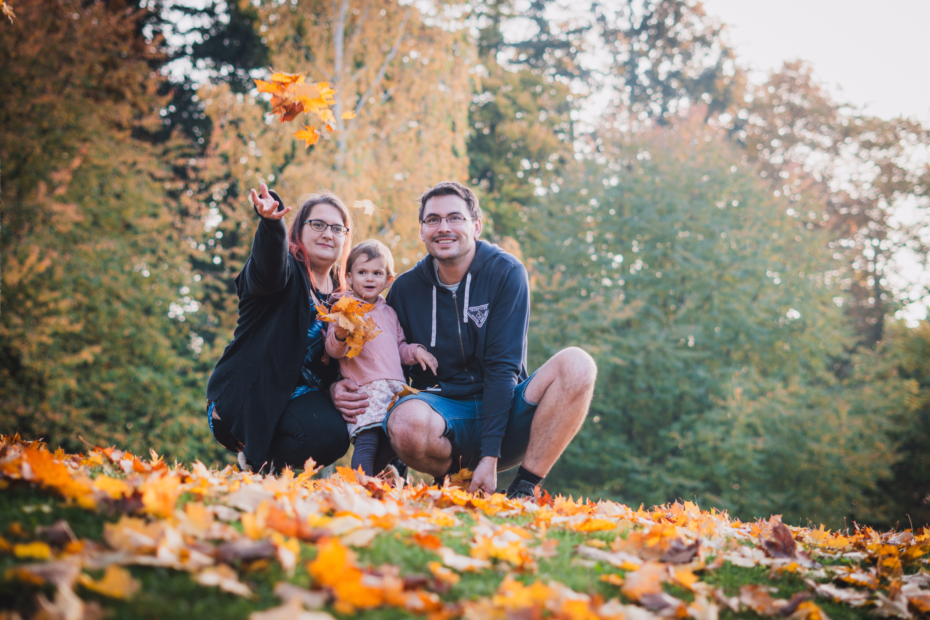 Šťastná rodina s malou dcerou se směje v podzimním listí během rodinného focení v parku.