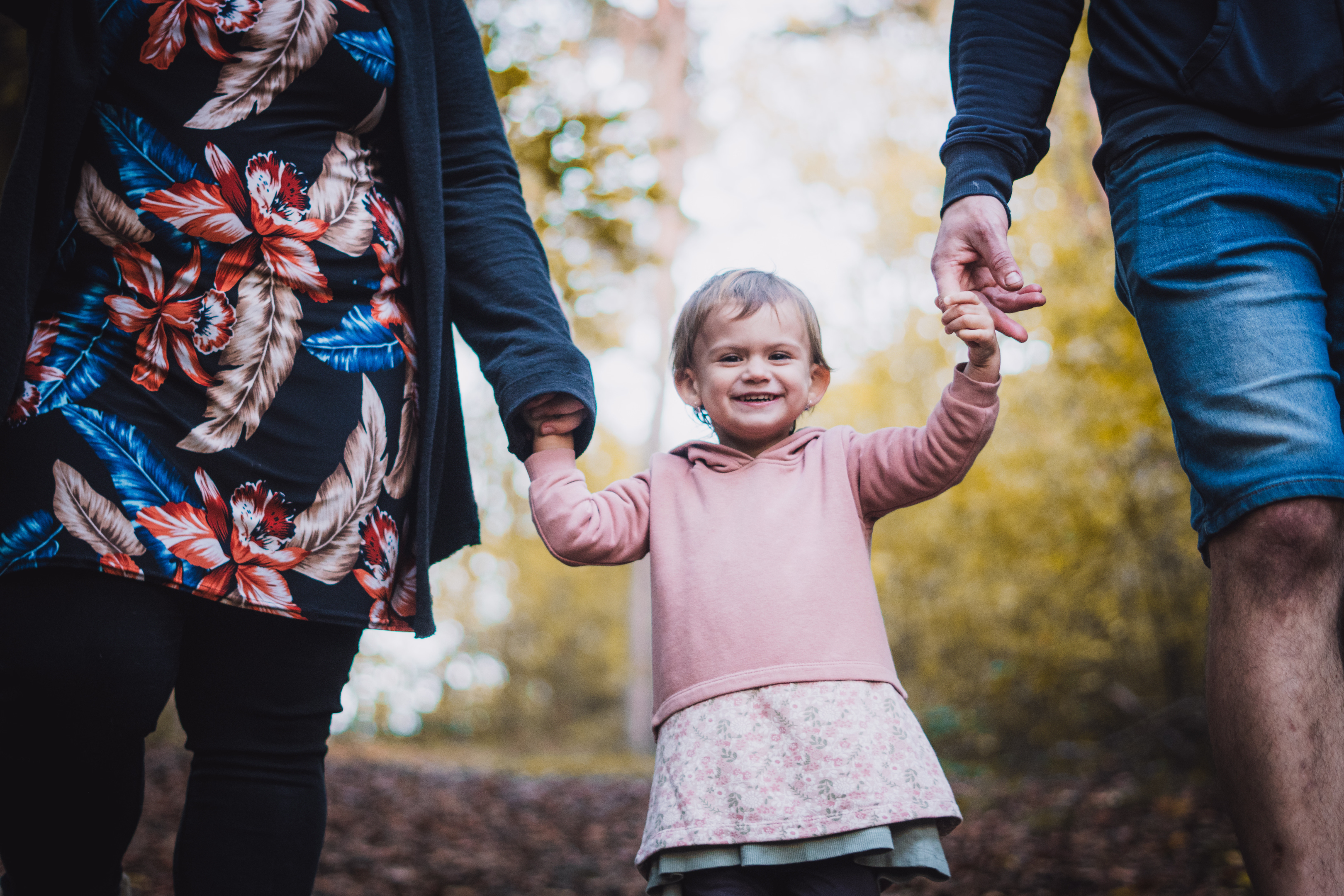 Usmívající se holčička drží své rodiče za ruce při procházce v podzimním lese, foceno z podhledu.
