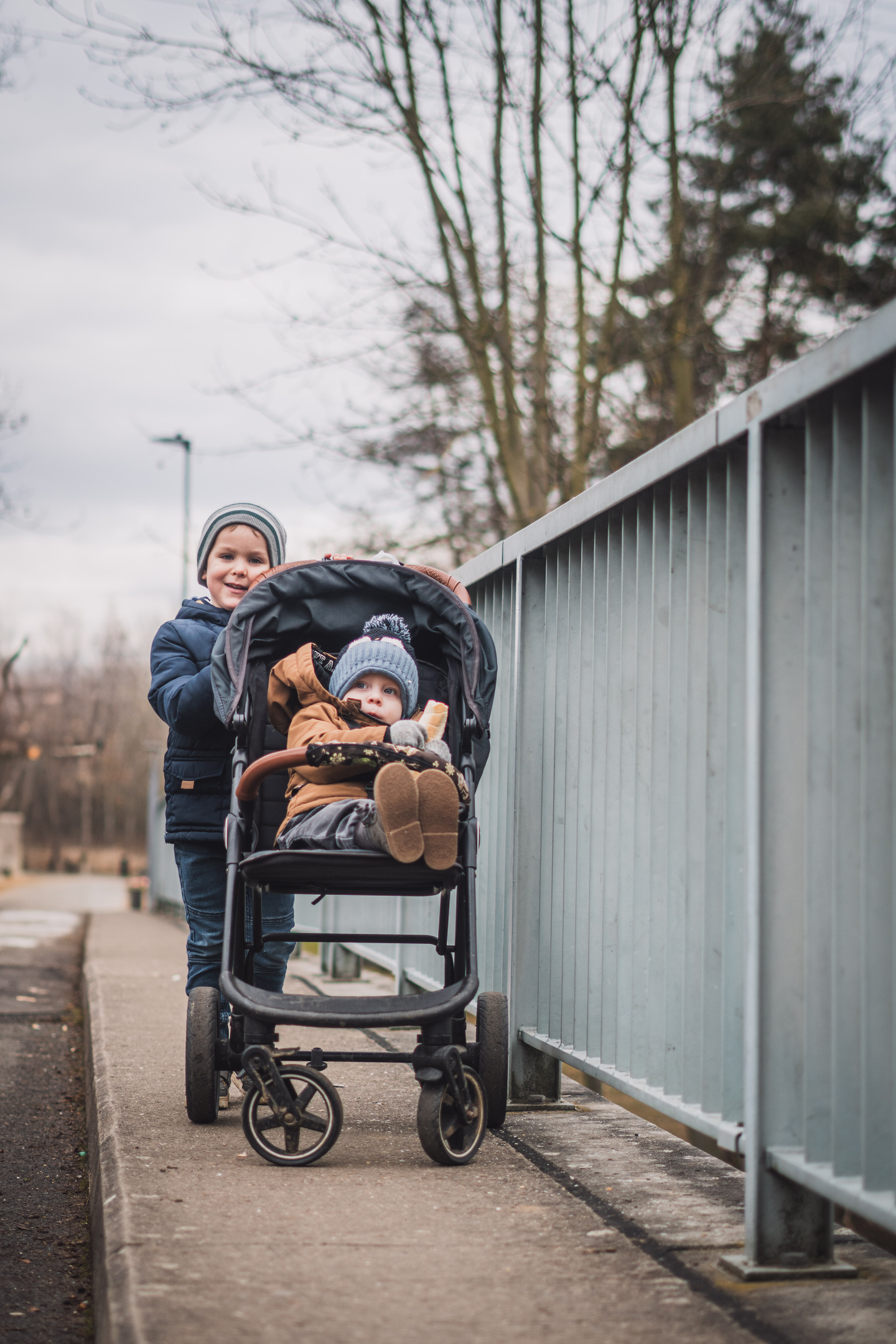 Portrét dvou bratrů – starší sourozenec se usmívá a tlačí kočárek s miminkem na chodníku.